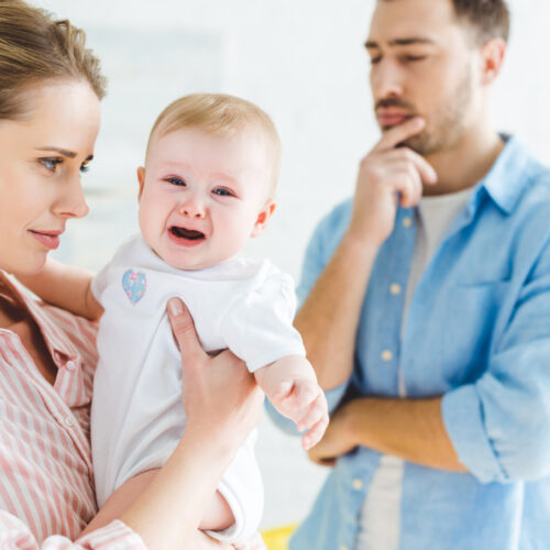Mother Holding Crying Infant Daughter On Hands And Thoughtful Father Standing Near